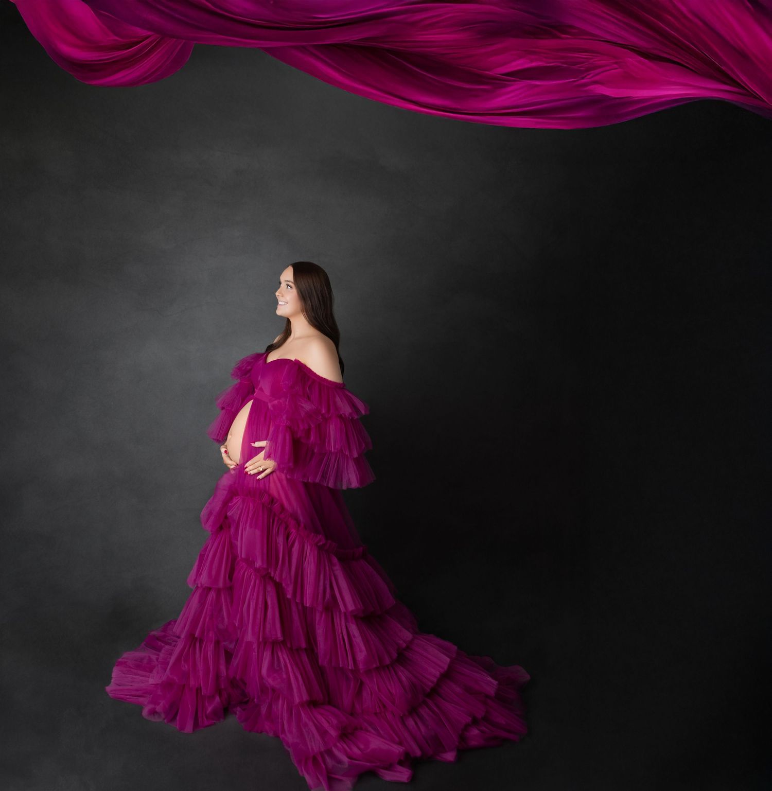 Expecting mother in magenta ruffled gown poses with flowing fabric against dramatic dark studio backdrop.
