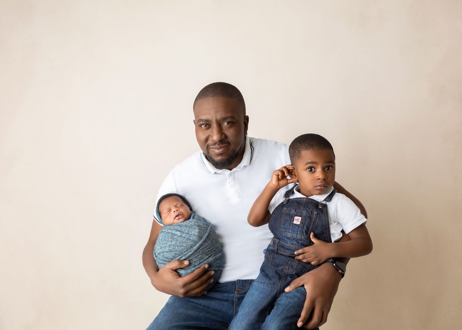 Father in white polo shirt holding swaddled newborn and toddler son in denim overalls in studio portrait.