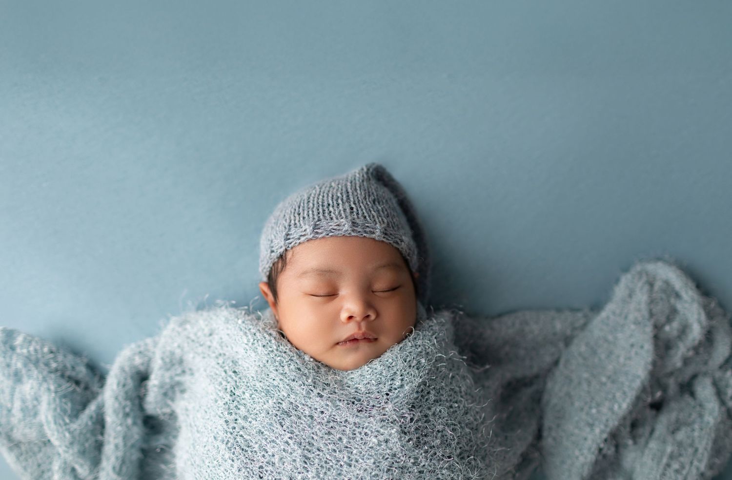 Peaceful newborn baby sleeping wrapped in a soft gray knit blanket and hat against a light blue background.