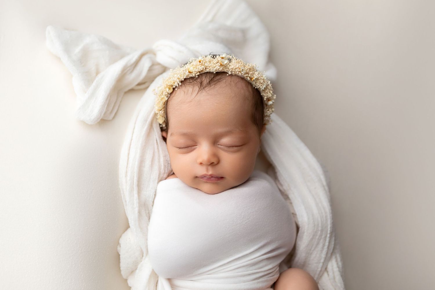 Sleeping newborn baby wrapped in white swaddle with floral headband on soft white background.