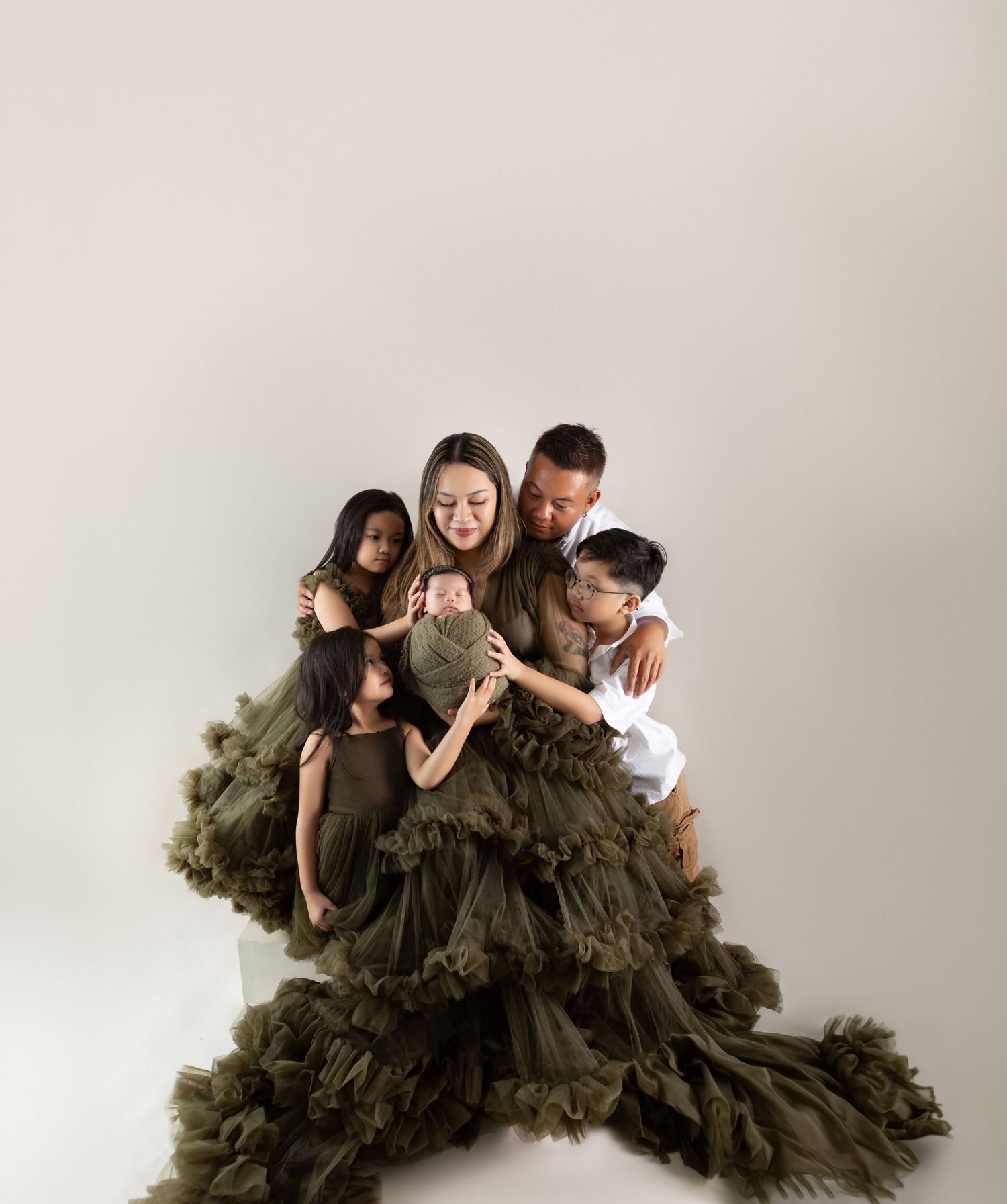 Family maternity photo with mother in olive tulle gown holding newborn, surrounded by father and three children in studio.
