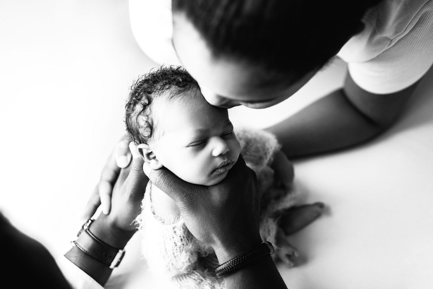 Black and white photo of a parent tenderly kissing a newborn baby while cradling them gently.