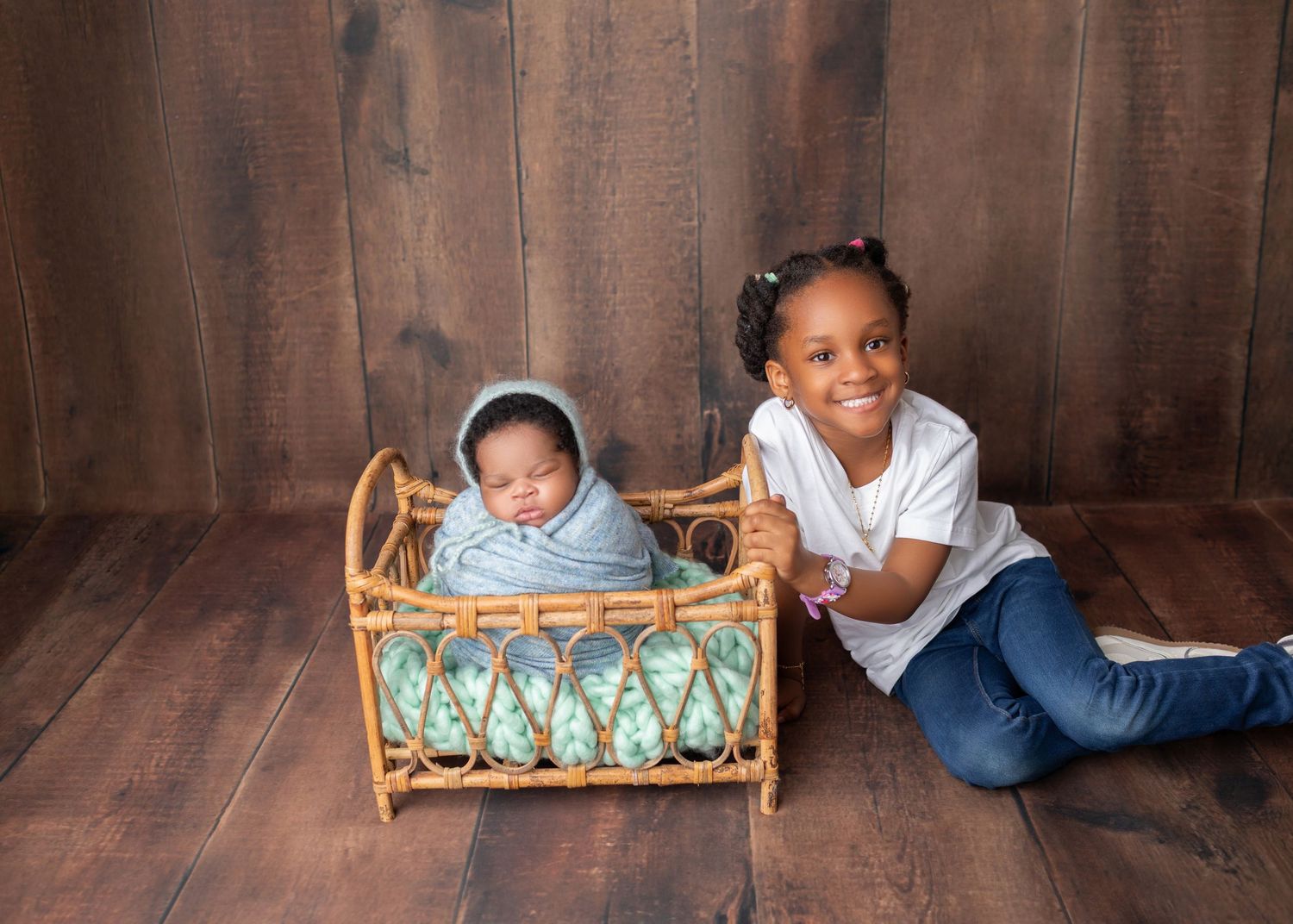 Young Black girl smiling beside newborn baby sibling in wicker cradle on wood floor.