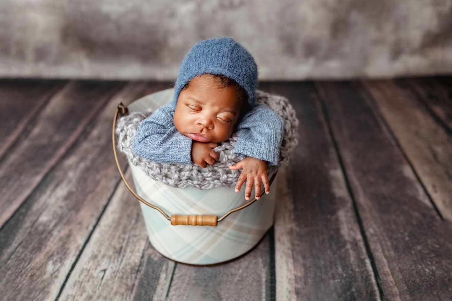 Sleeping newborn baby in blue knit hat posed in decorative bucket on rustic wood floor.