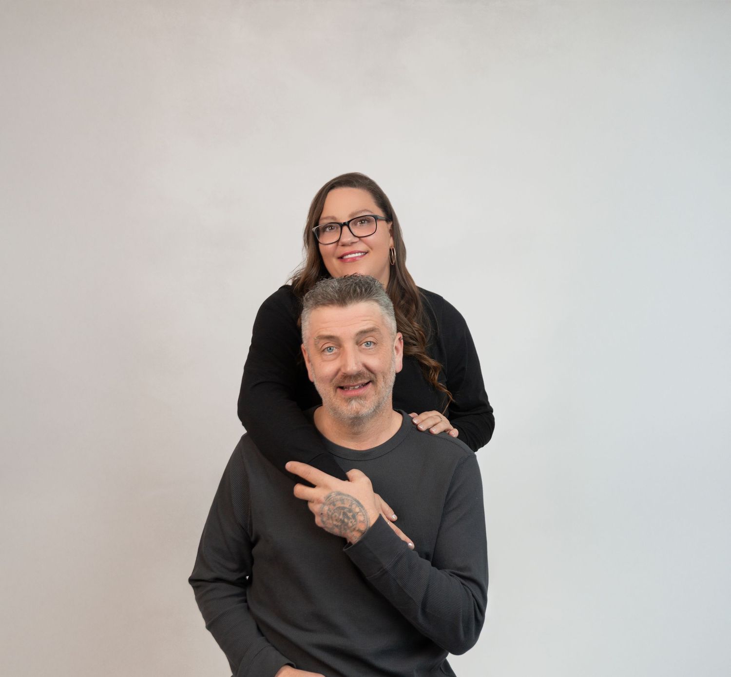 A woman rests her chin on a tattooed mans shoulder as they pose together against a white background.