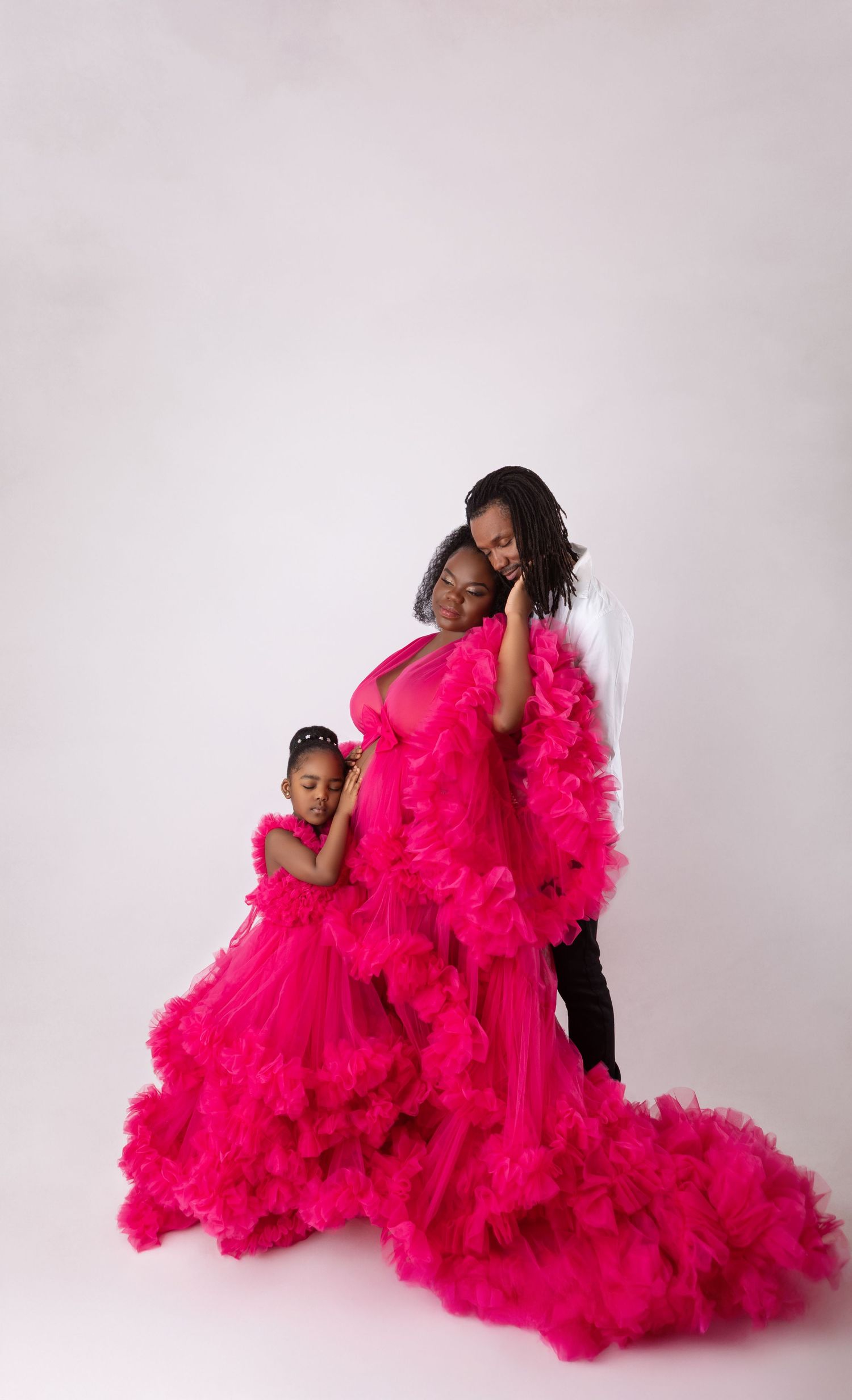 Pregnant woman and family in matching hot pink tulle gowns embrace in soft white studio.