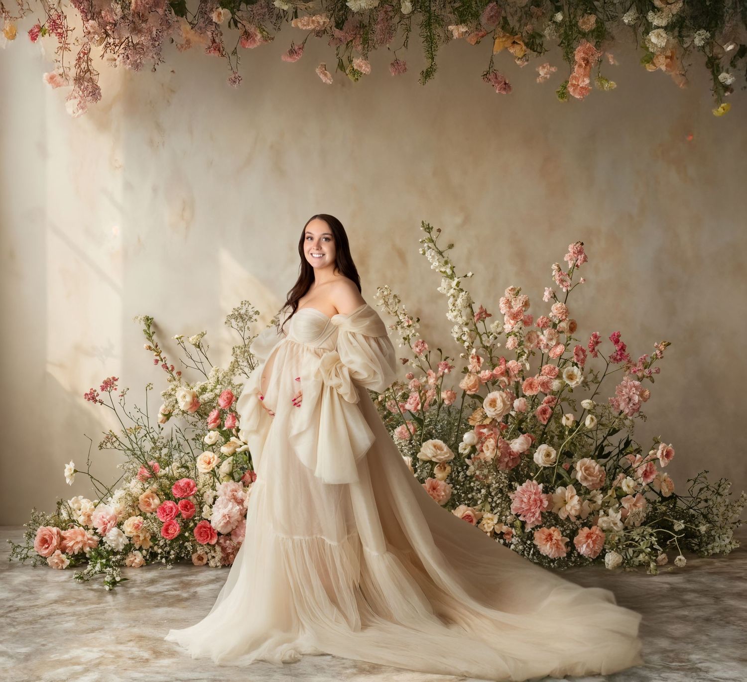 Pregnant woman in stunning cream off-shoulder tulle gown surrounded by lush pink and white floral arrangements in studio.