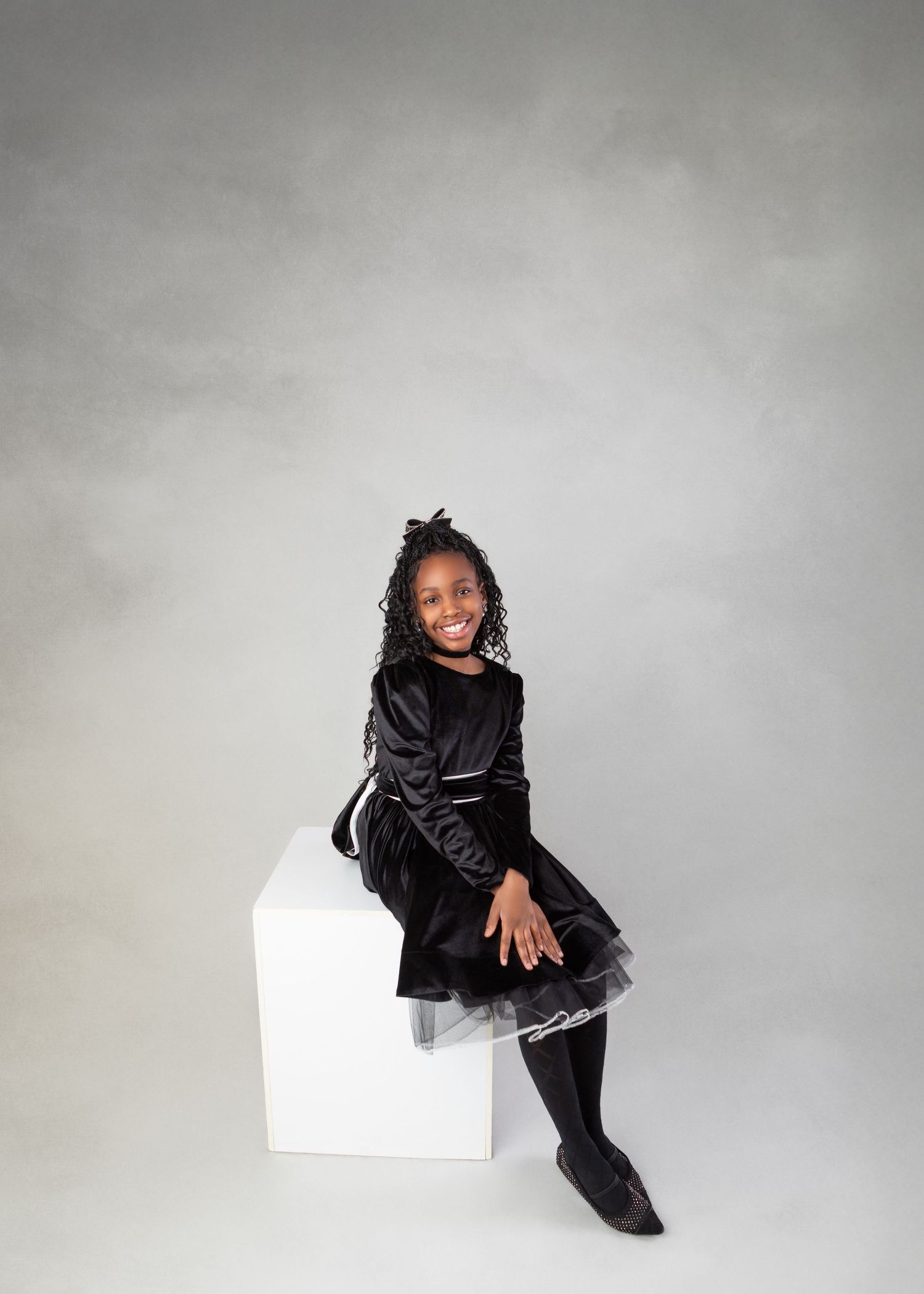 Young girl in black velvet dress sits smiling on white cube against gray background.