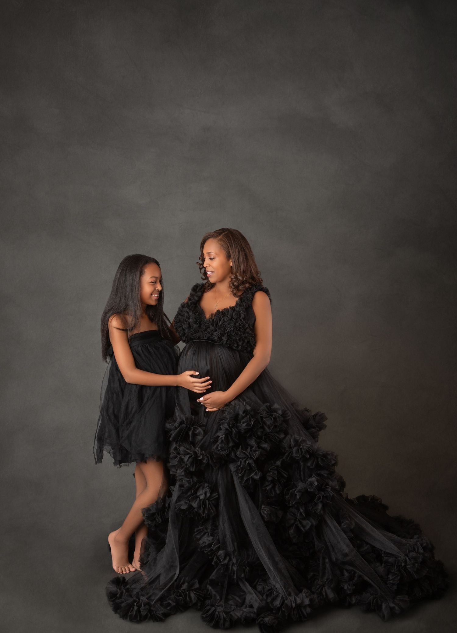 Pregnant woman in black ruffled gown poses with young girl in black dress for elegant maternity studio portrait.