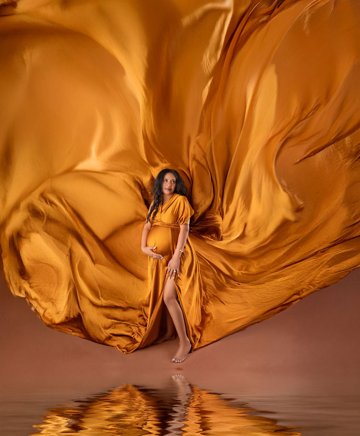 Pregnant woman in flowing golden orange gown with dramatic fabric billowing, reflected in water below.