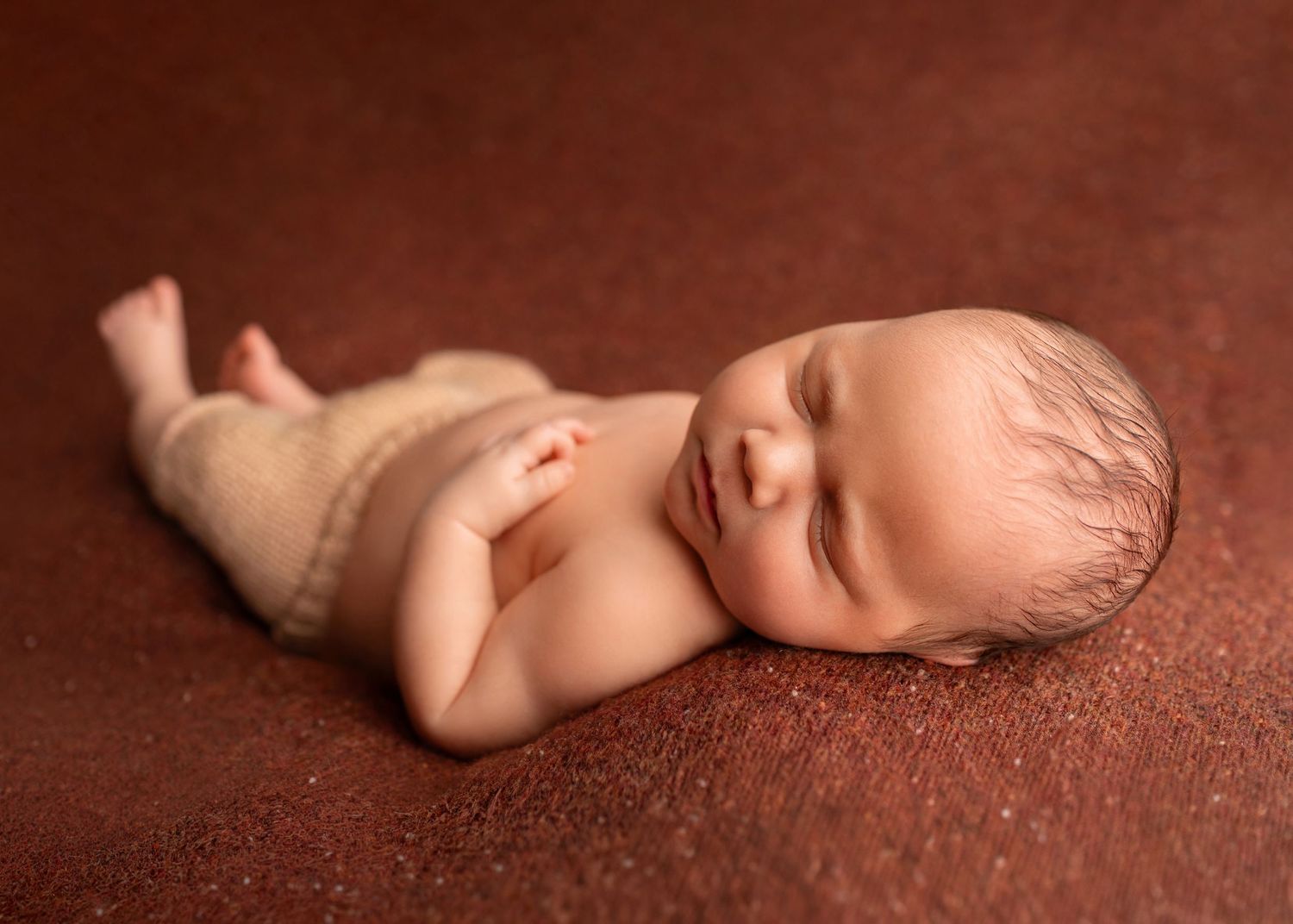 Peaceful newborn baby sleeping on a brown textured backdrop, wearing a knit wrap, smiling softly.