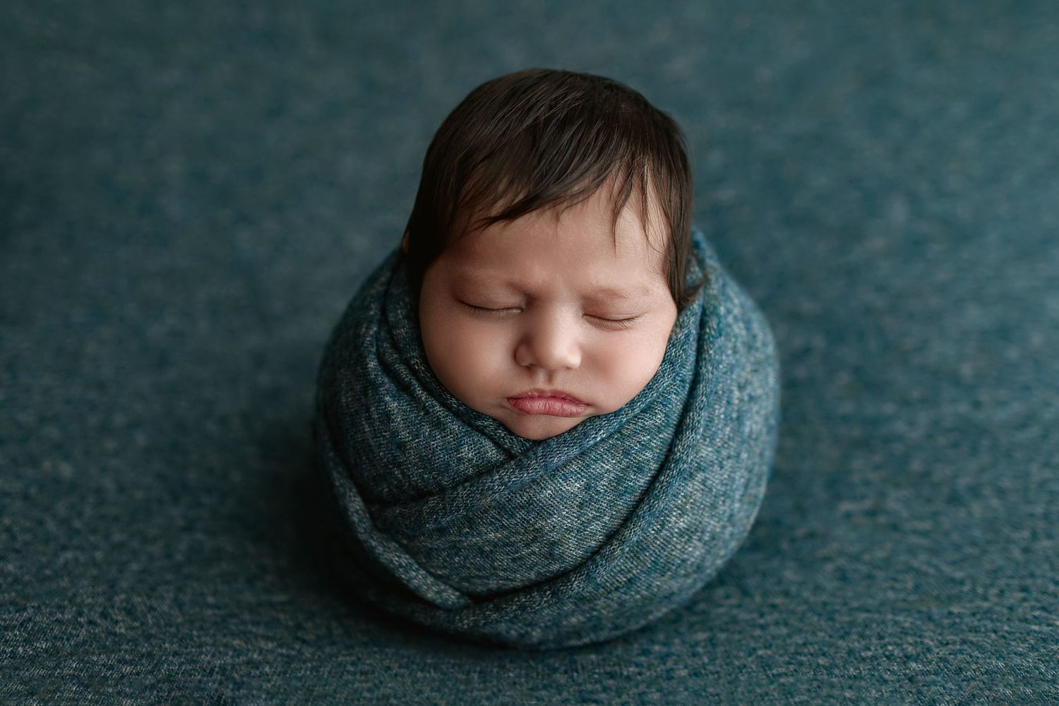 Newborn baby peacefully sleeping, swaddled in a teal wrap, posed in a potato sack position on a teal backdrop.