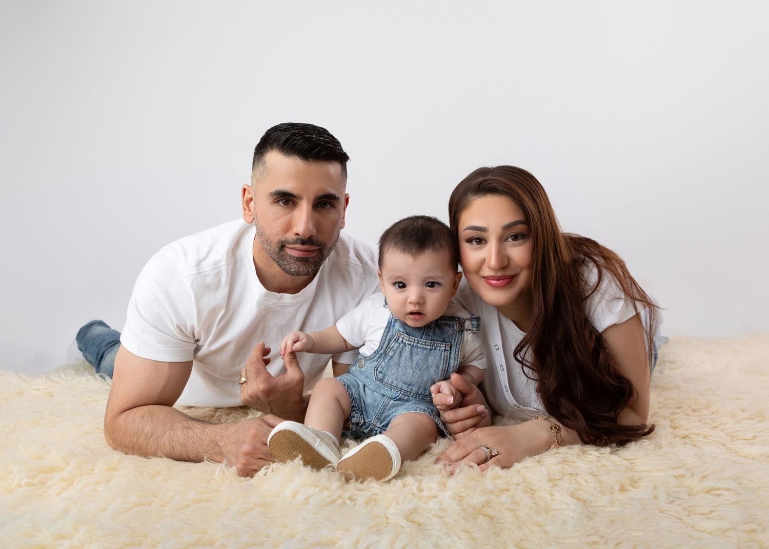 A young family with a baby boy poses on a fluffy white rug, wearing casual white outfits in a bright studio.