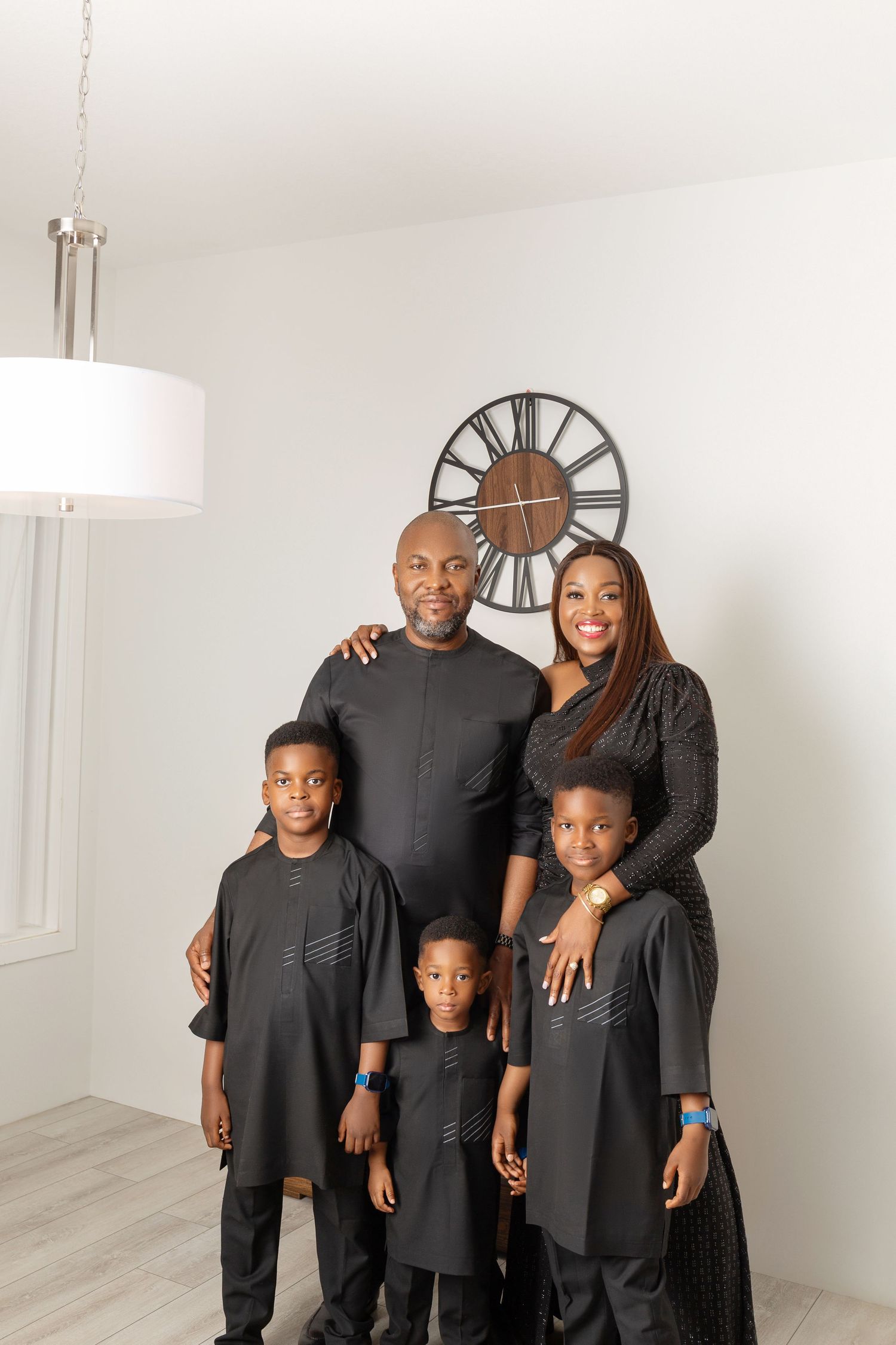 Family of five in matching black outfits posing together in a bright white room with a decorative wall clock.