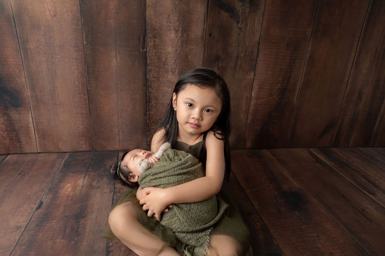 Big sister in green romper gently cradles wrapped newborn baby against wooden backdrop.
