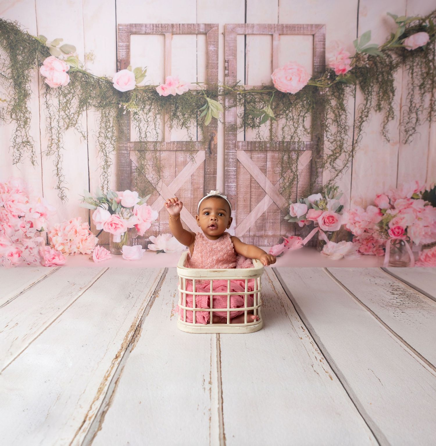 Cute baby girl in pink outfit sitting in white basket with floral garland barn backdrop.