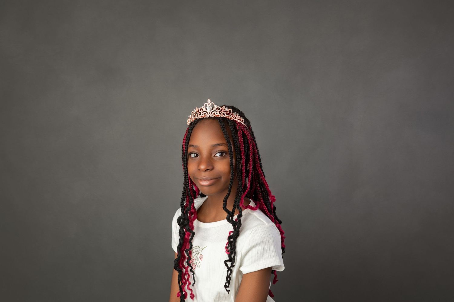 Young Black girl with red braided hair wearing a tiara and white shirt, posing against a gray background.