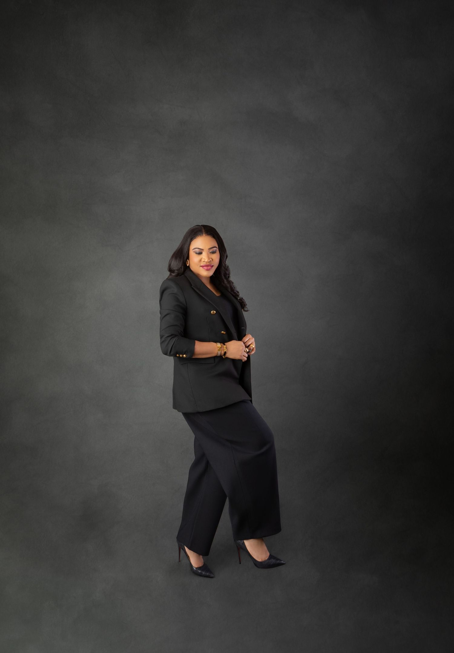 Woman in black blazer and wide-leg pants poses confidently against a dark gray studio background.