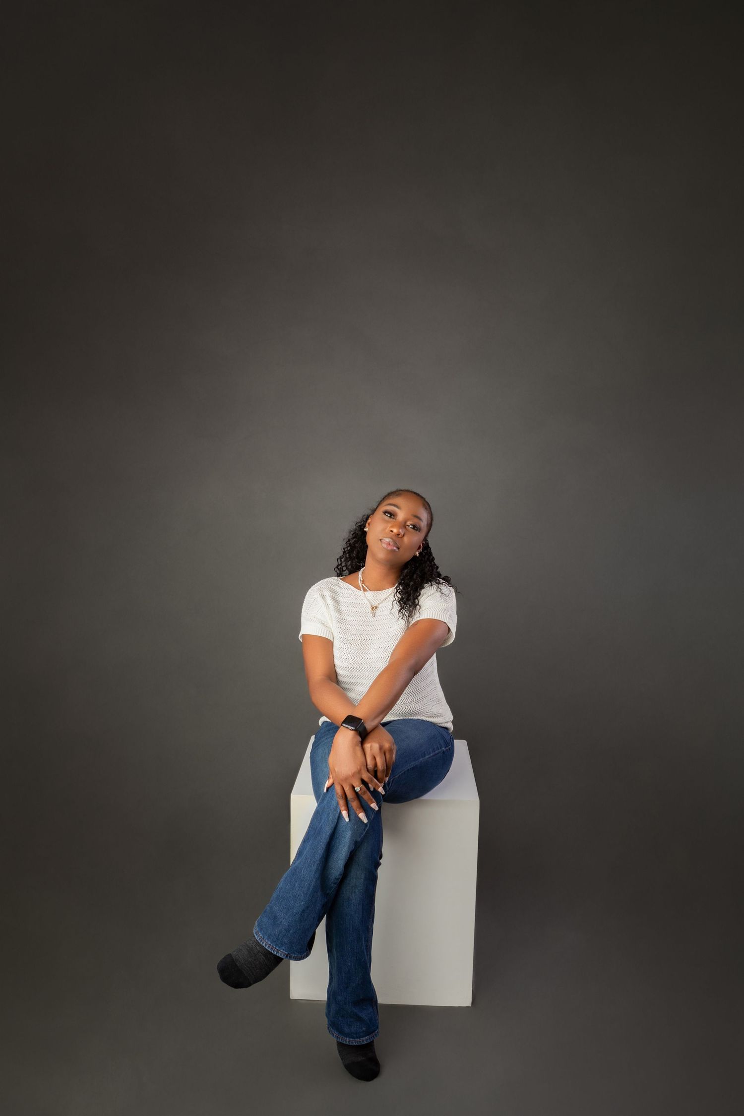 Young woman in white t-shirt and blue jeans sitting on white cube against gray studio background.