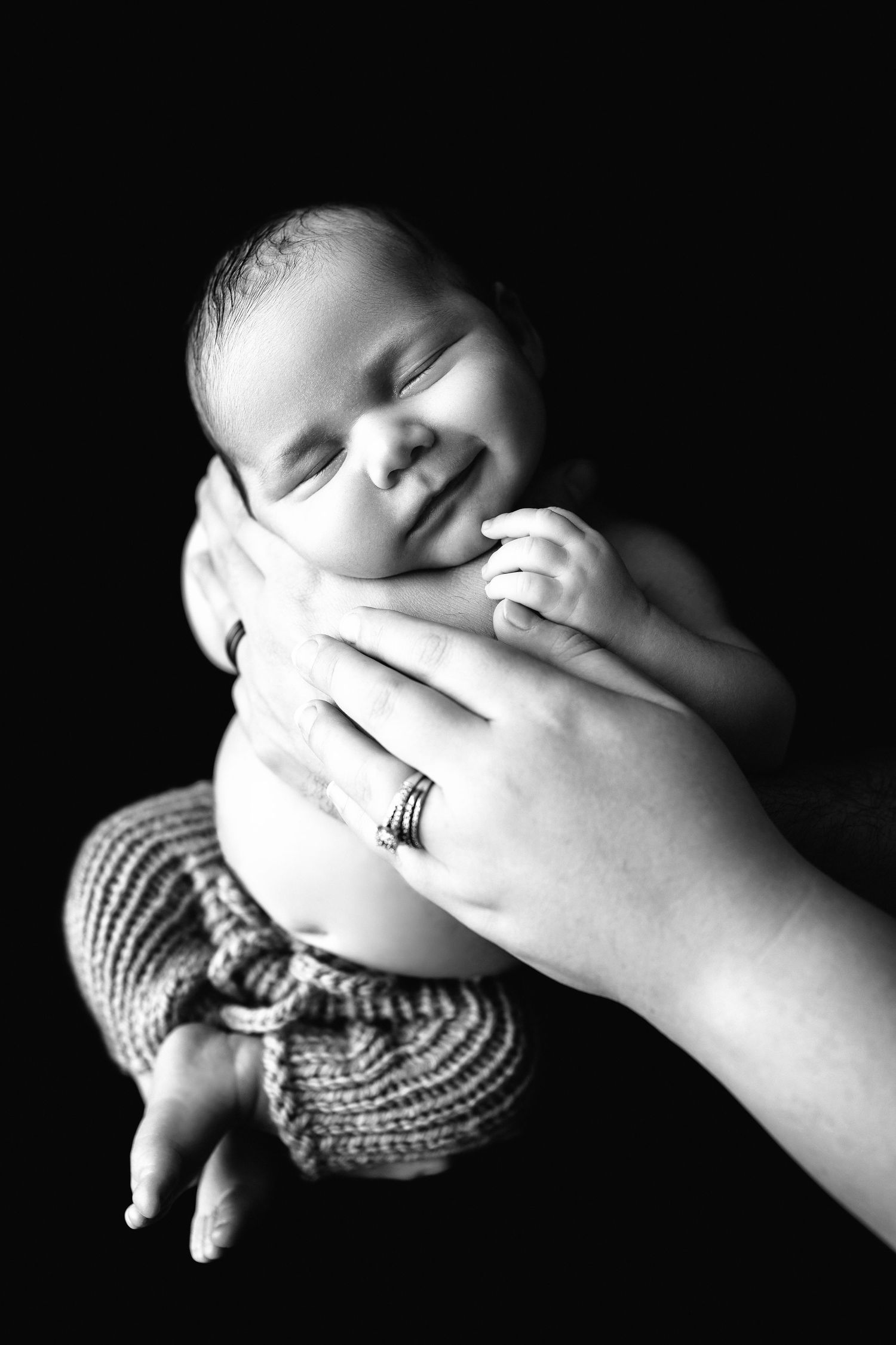 Black and white portrait of smiling newborn baby cradled in parents hands against dark background.
