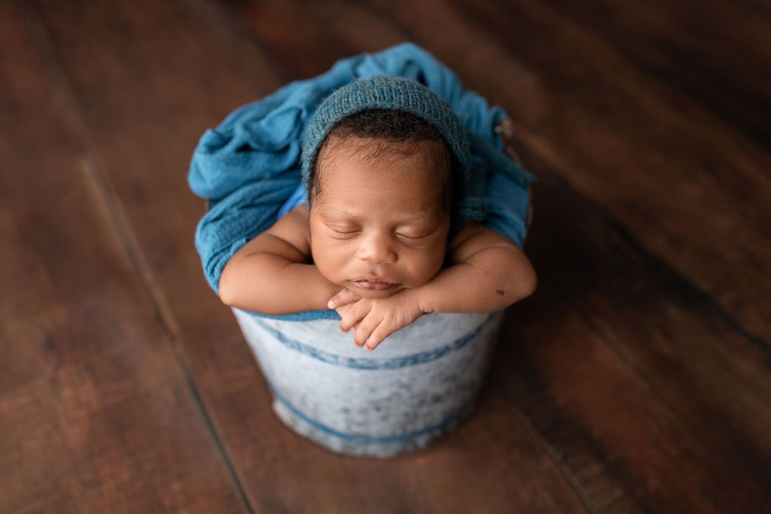 Sleeping newborn baby wrapped in blue fabric resting chin on hands inside a rustic bucket on wood floor.