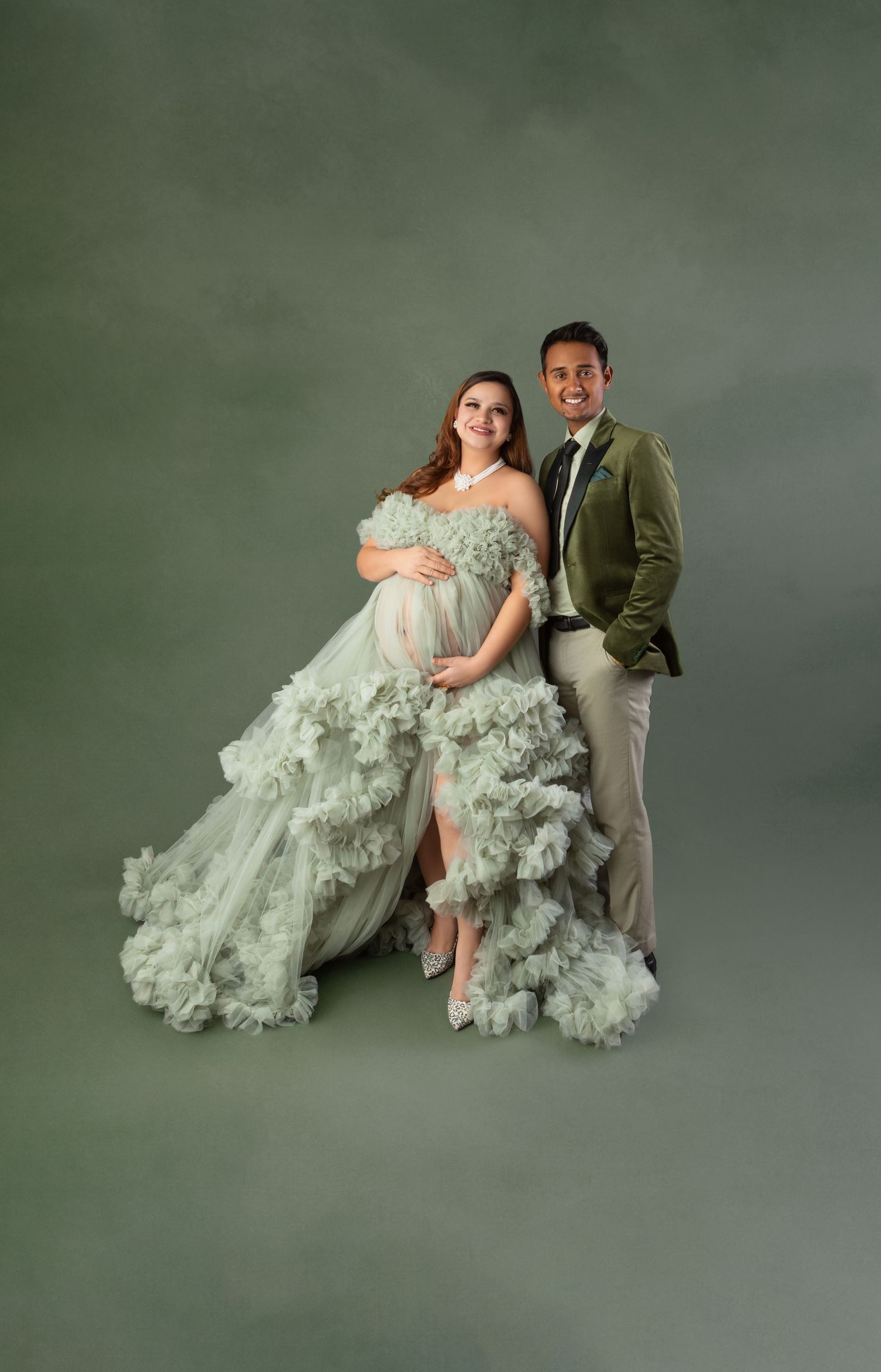 Expecting couple posing together in a studio, woman in sage green ruffled maternity gown.