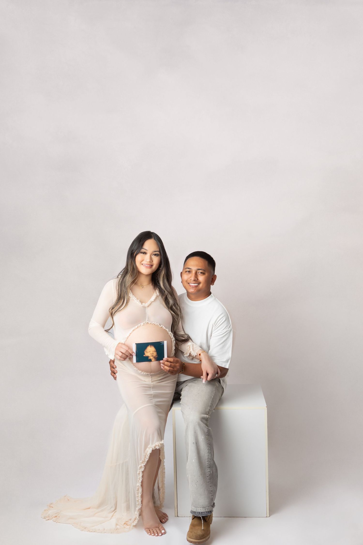 Pregnant couple smiling and holding ultrasound photo in a bright studio maternity photoshoot.