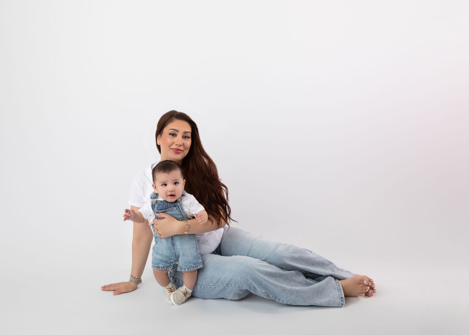 Mother sitting on floor holding baby in denim overalls against white background.