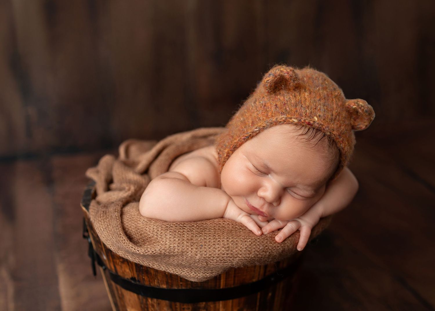 Peaceful newborn wearing brown bear ear bonnet sleeping in wooden barrel with tan wrap.