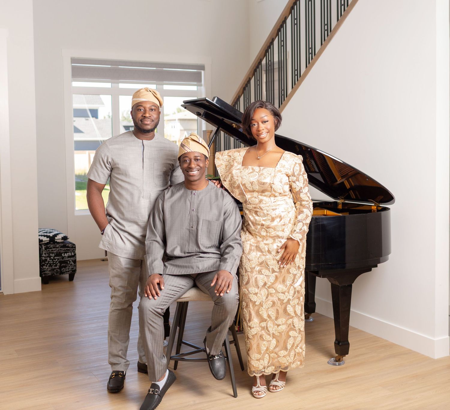 Three people in traditional African attire pose together near a grand piano and staircase in an elegant home.