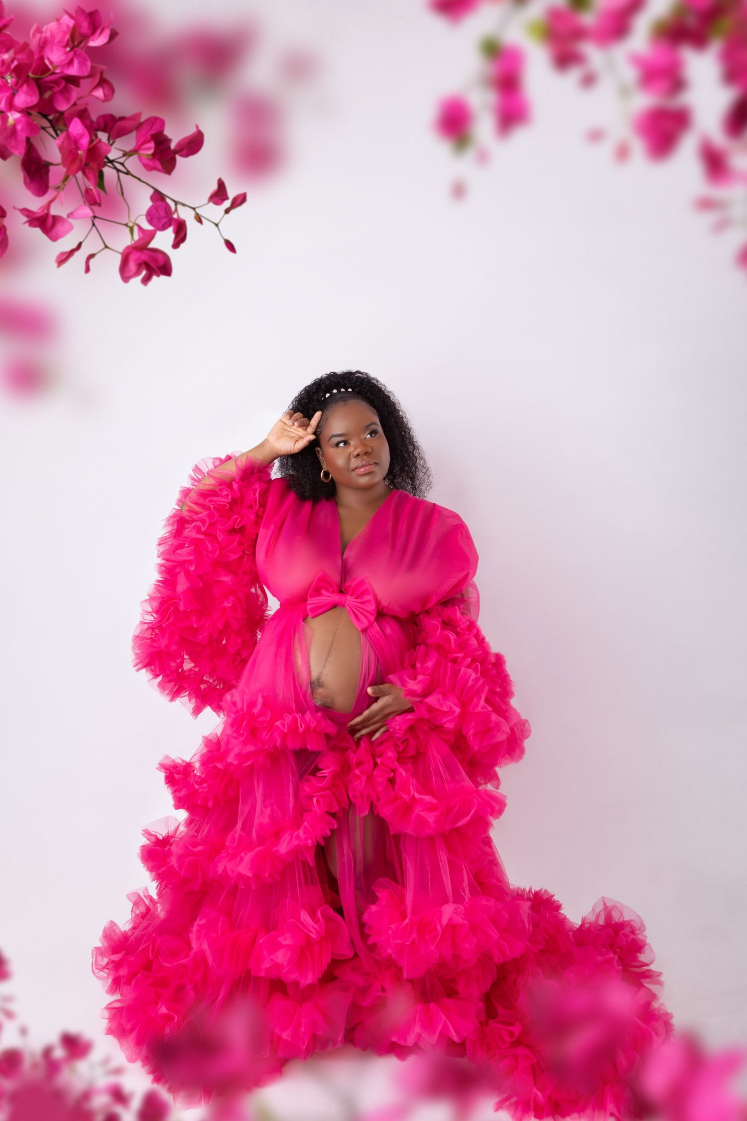 A pregnant woman in a vibrant pink ruffled maternity gown poses among pink bougainvillea flowers.