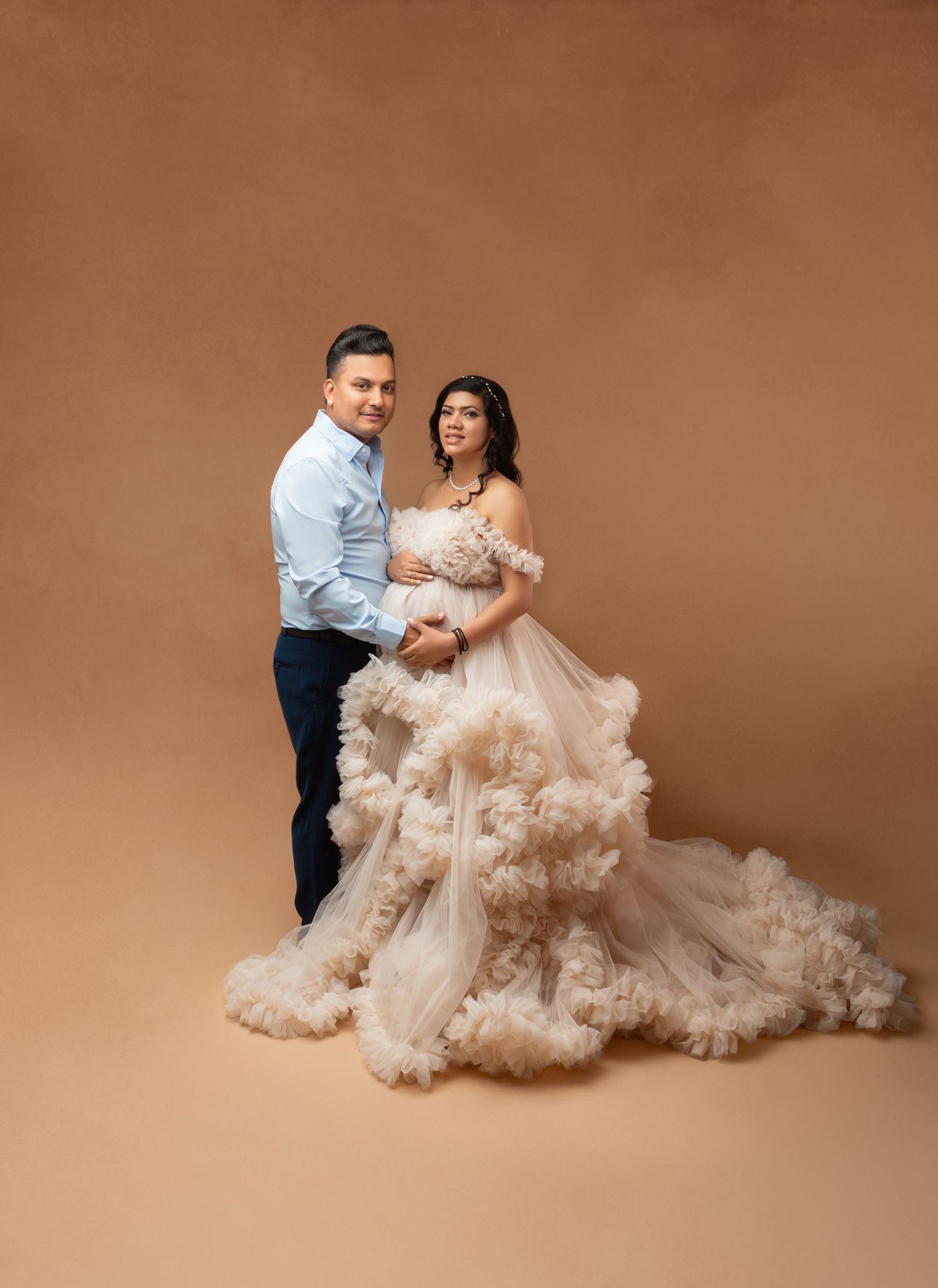 Couple posing for maternity photo shoot, woman in elegant ruffled tulle gown, man in light blue shirt, warm beige backdrop.