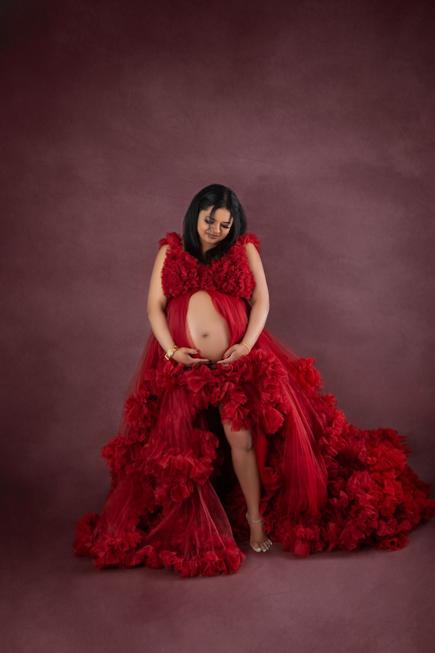 Pregnant woman in elegant red ruffled maternity gown cradling baby bump against dark rose studio backdrop.