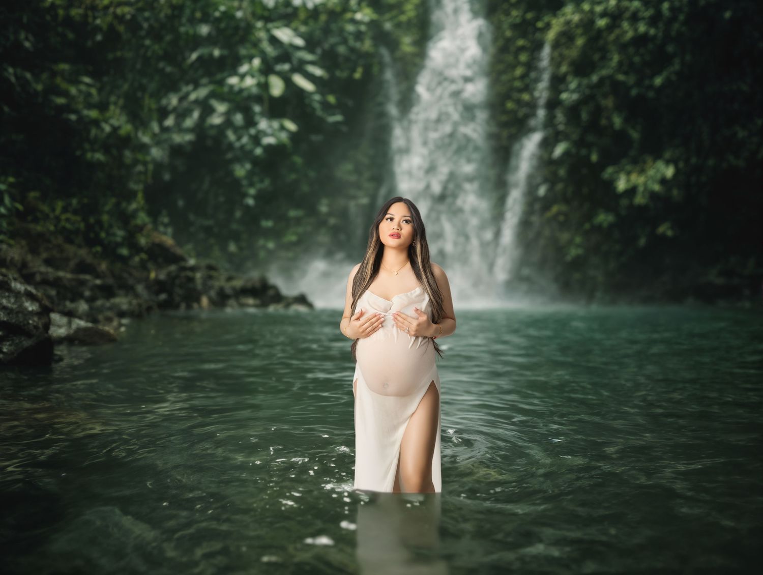 Pregnant woman in white dress standing in turquoise water before a lush green waterfall during maternity photoshoot.