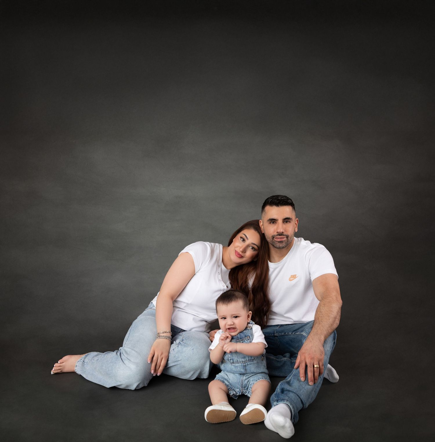 A young family with a baby sits on a studio floor, wearing white shirts and jeans, posing for a portrait.