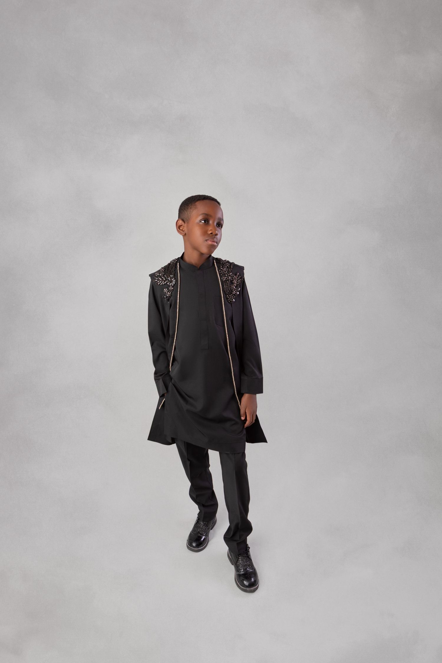Young boy in black embellished vest, tunic, and trousers posing against gray background.