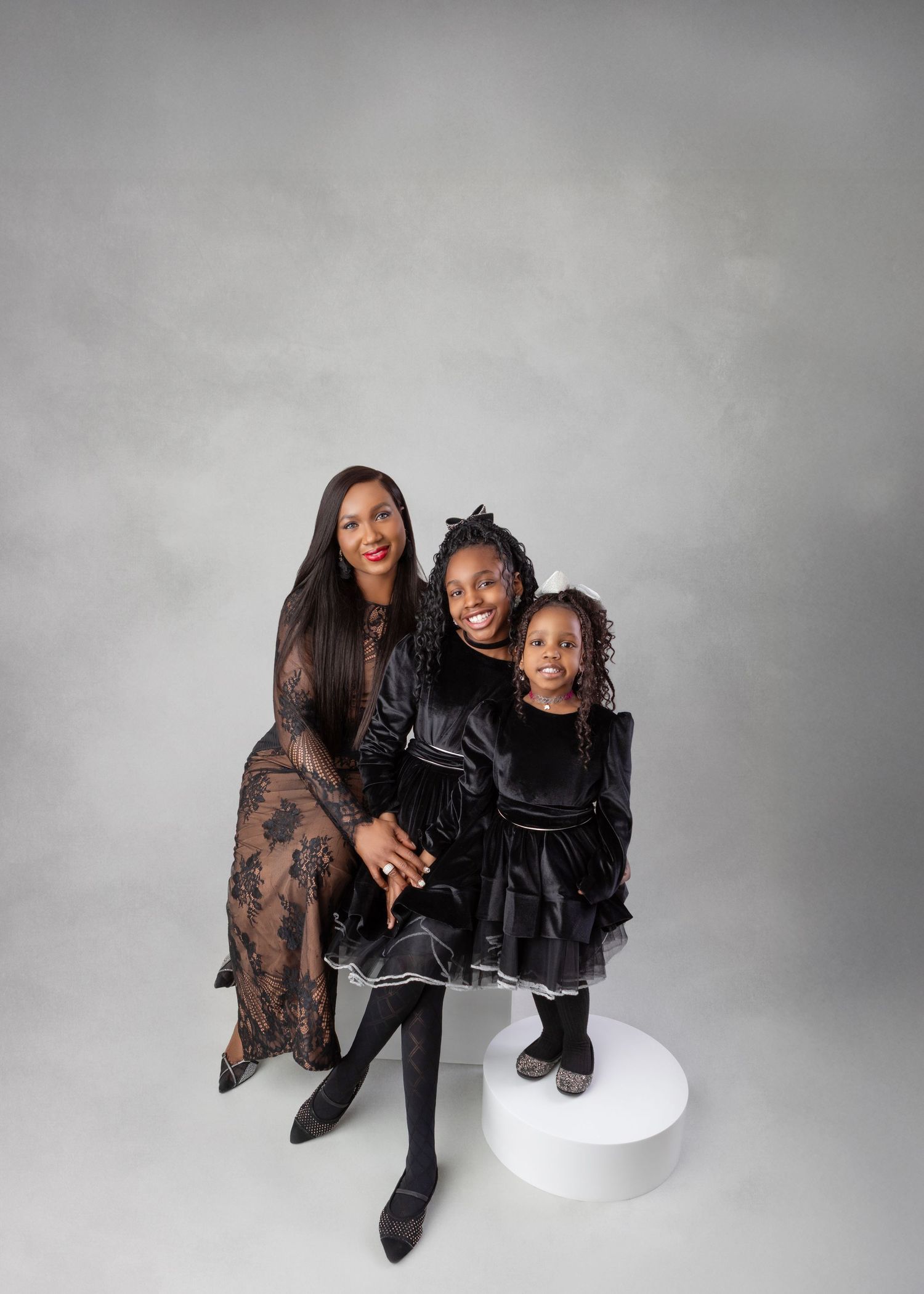 A woman and two young girls pose together in matching black outfits against a gray studio background.