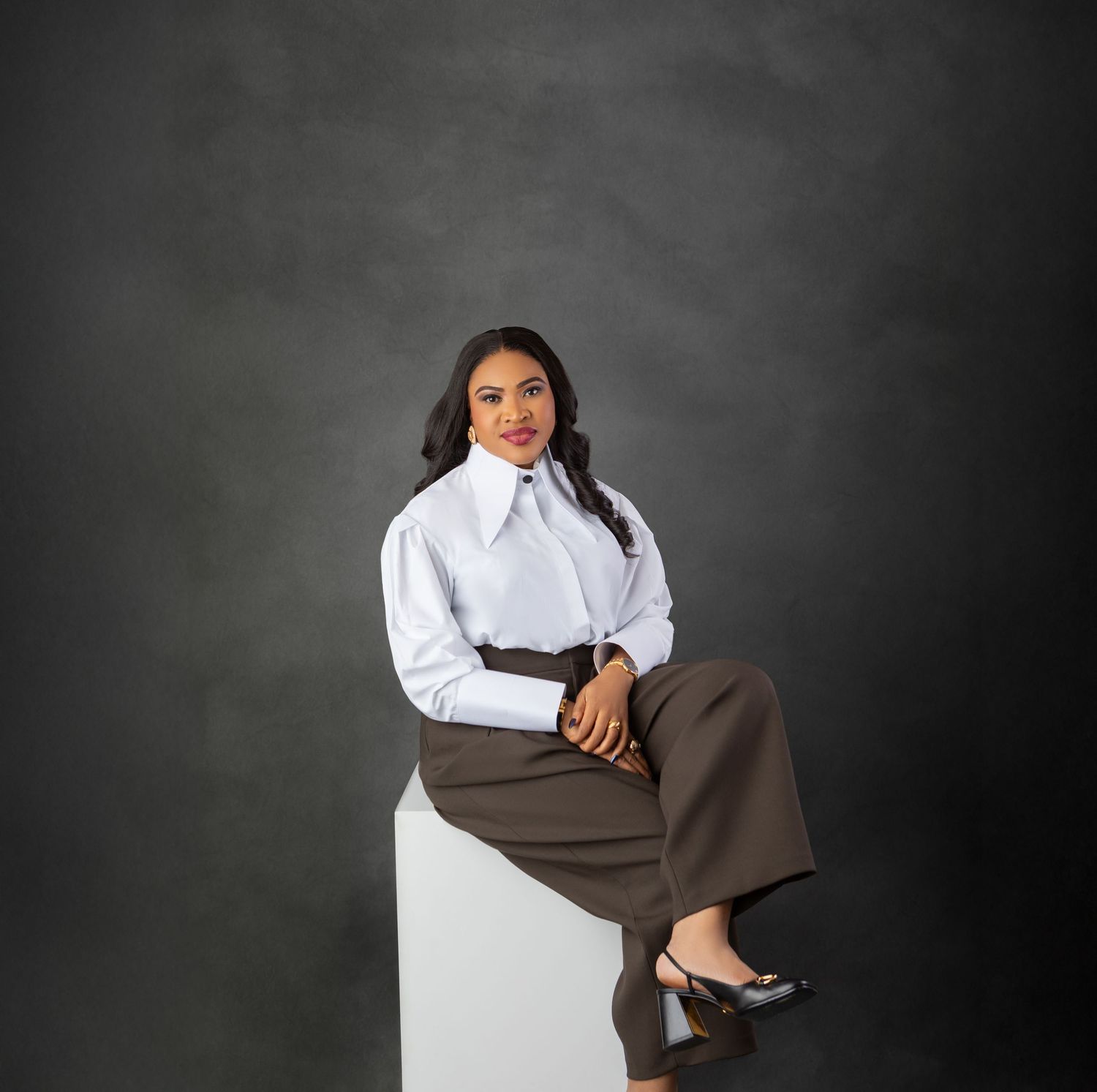 Woman in white blouse and brown wide-leg trousers sits confidently on white cube against dark gray backdrop.
