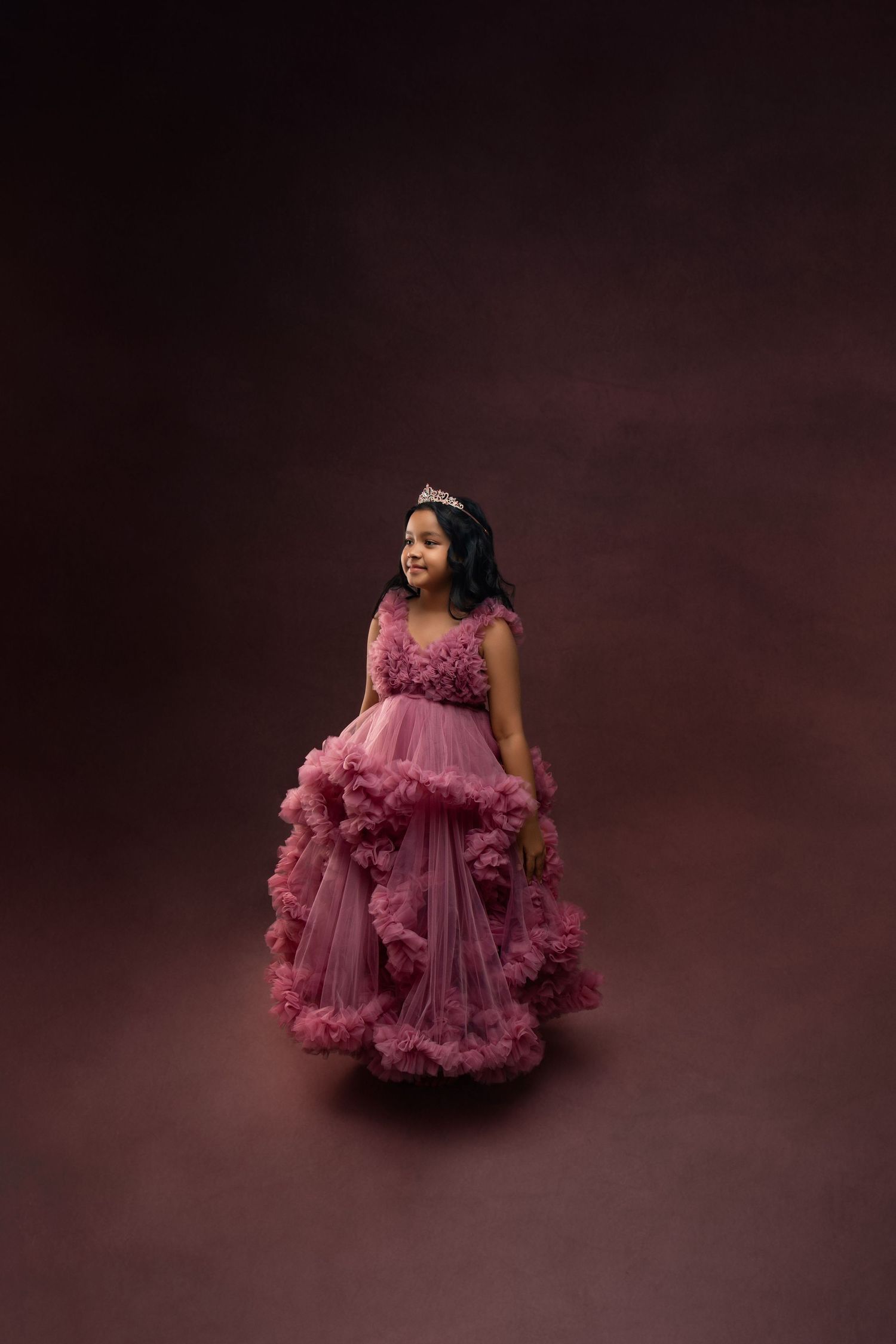 Young girl in a mauve ruffled tulle ball gown and tiara posing in a professional photo studio.