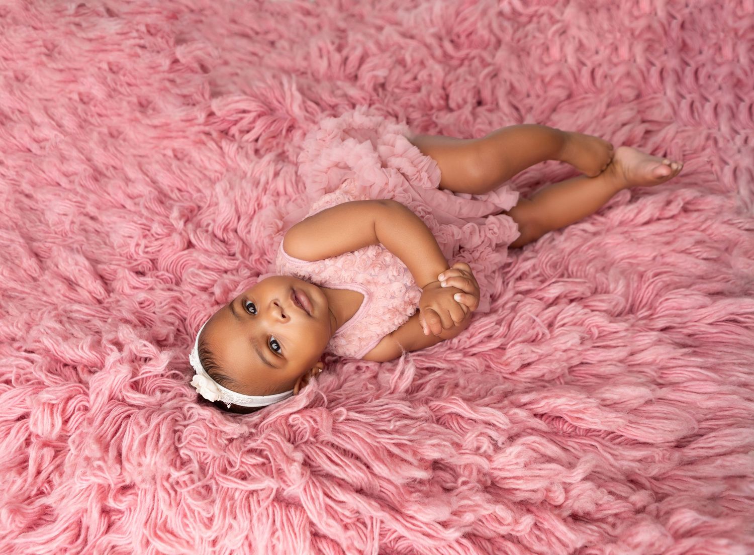Baby girl in pink romper lying on a fluffy pink rug during a newborn photo session.