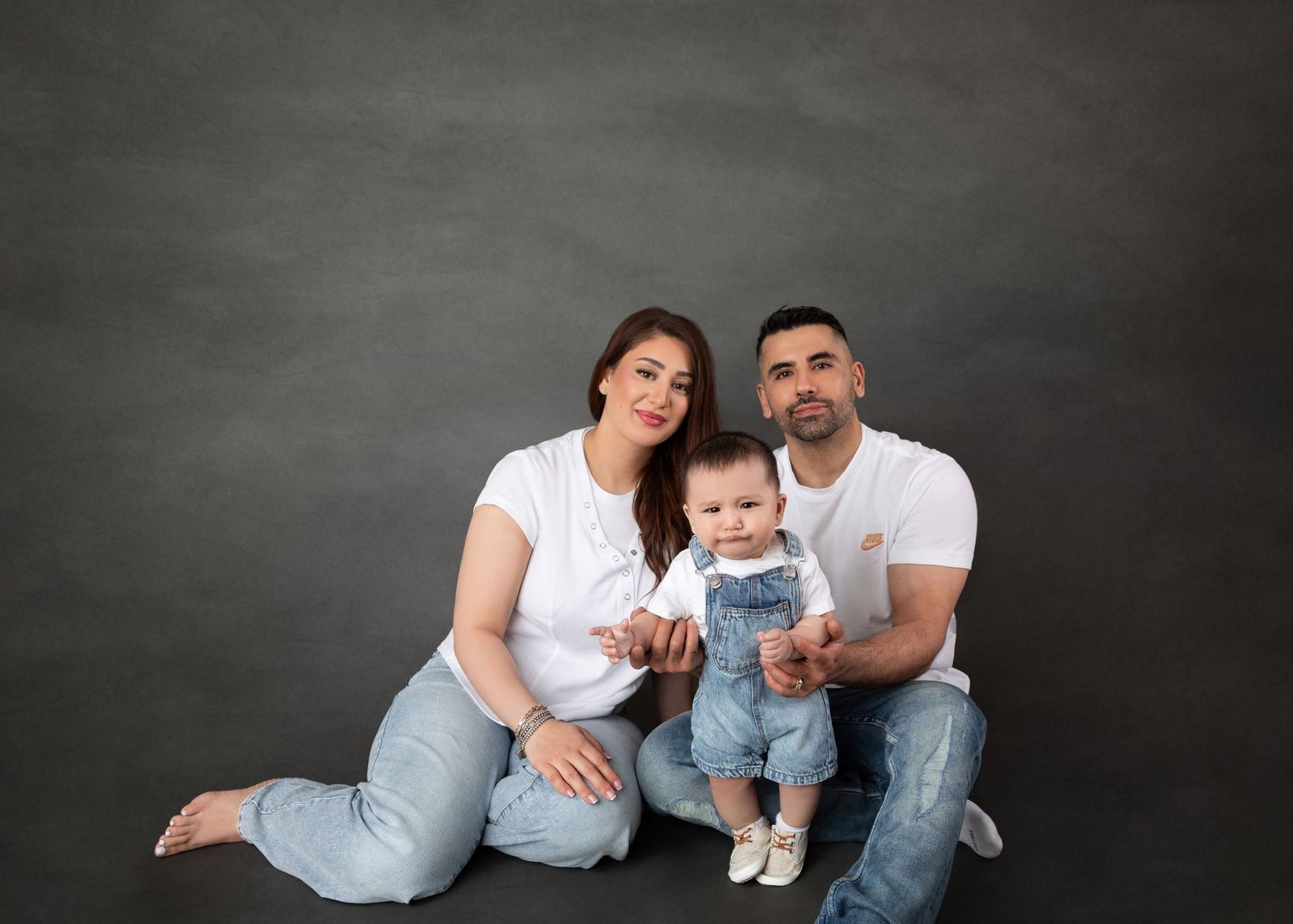 Parents posing with baby in denim overalls seated on grey studio floor.