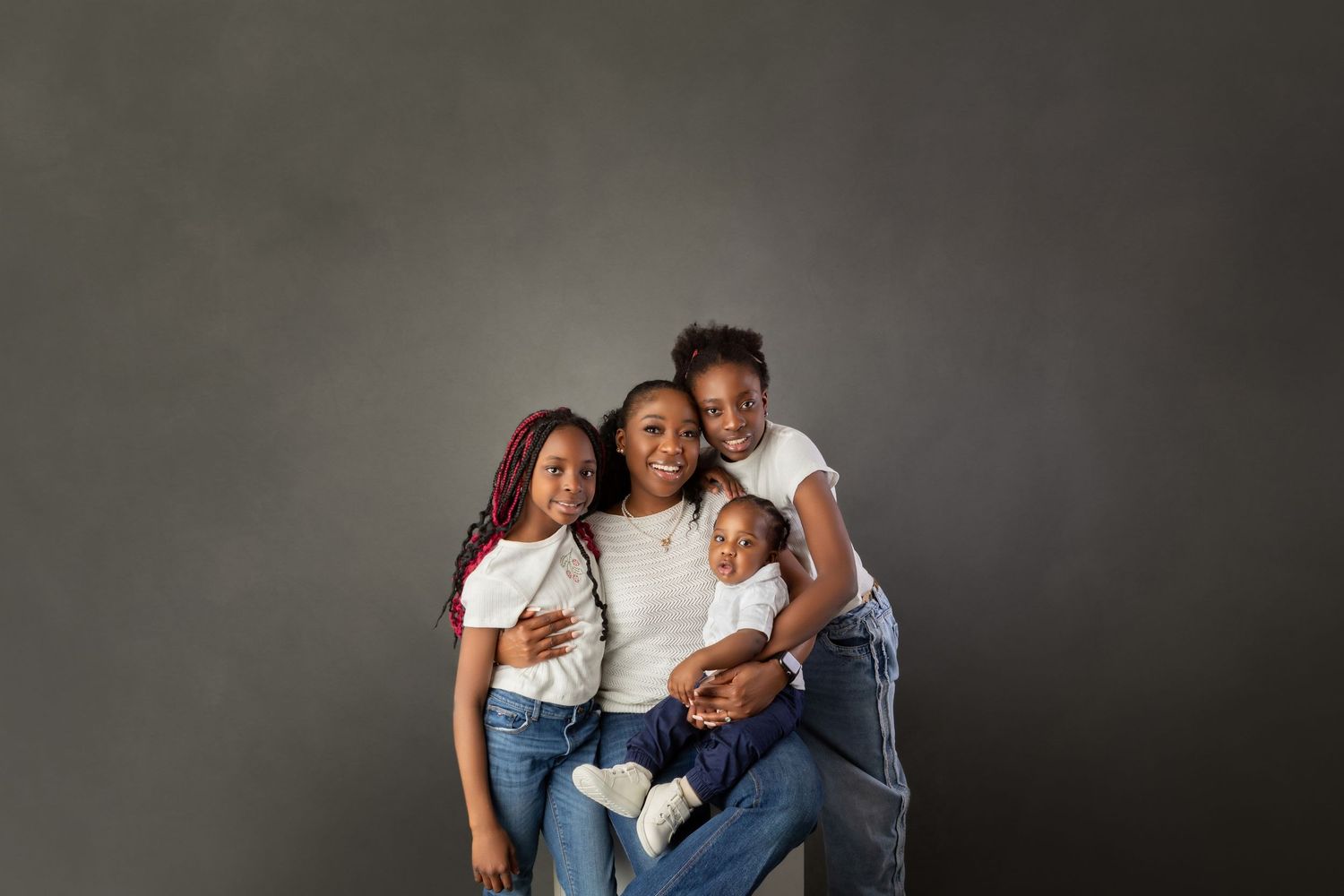 Black mother sitting with three children in white shirts and jeans smiling in studio portrait.