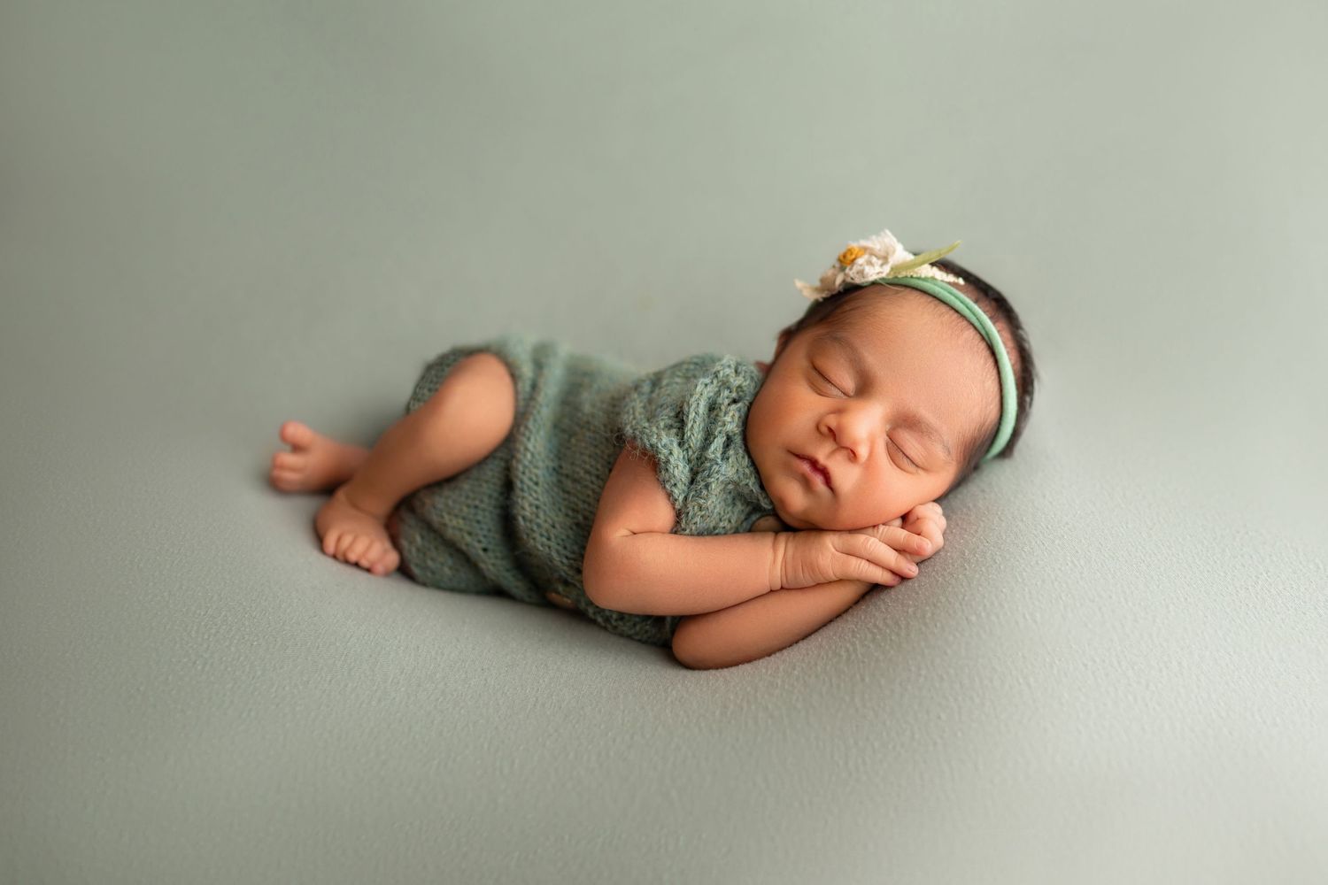 Newborn baby sleeping peacefully in sage green knit outfit with flower headband on a soft green backdrop.