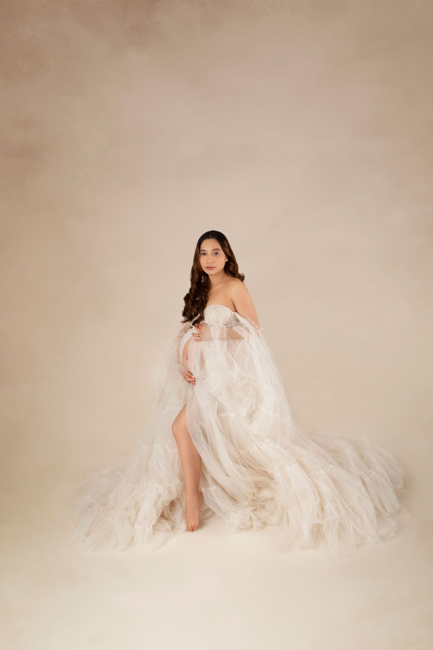 A pregnant woman in a sheer white ruffled tulle gown poses gracefully against a warm beige backdrop.