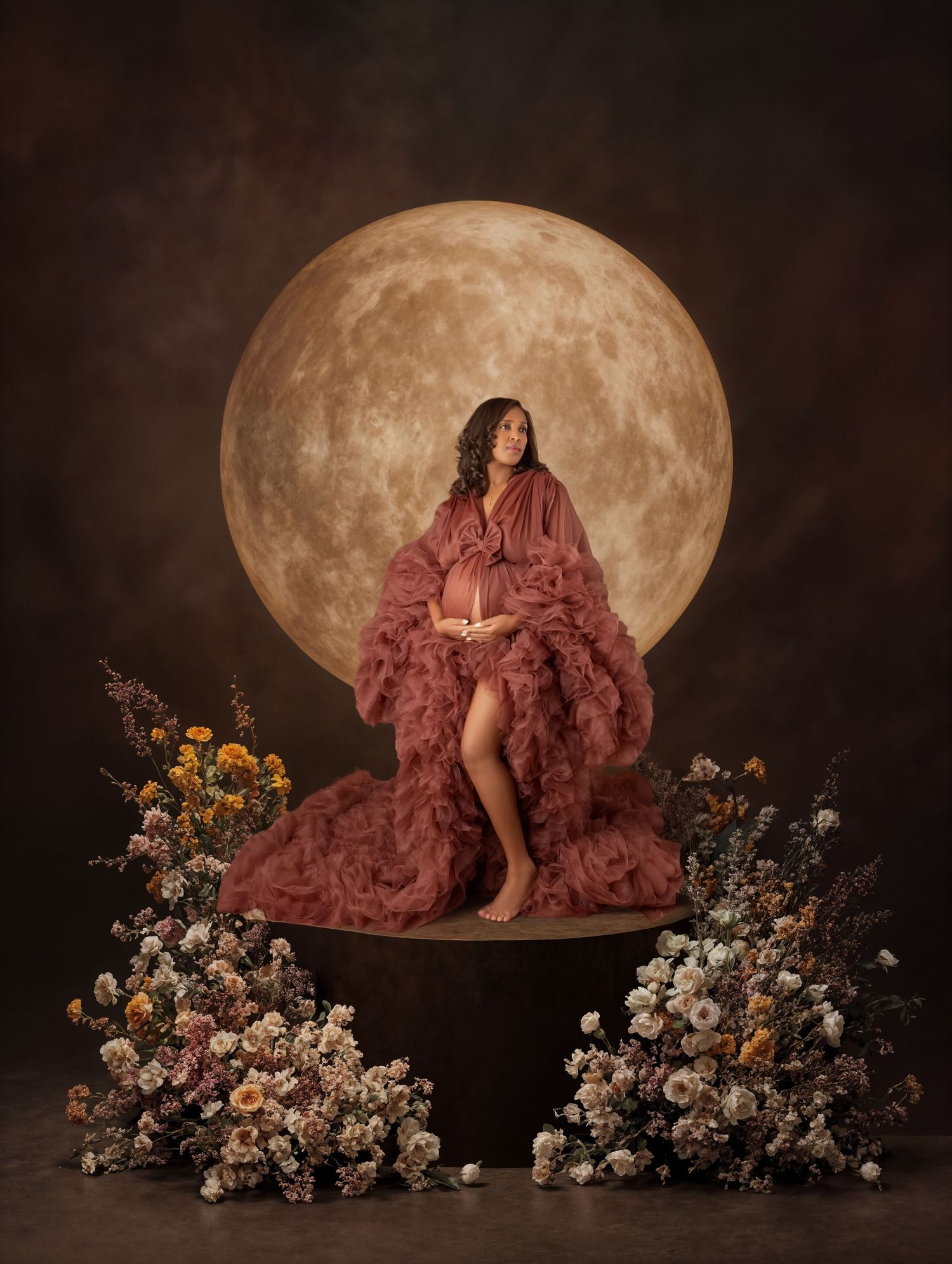Pregnant woman in dusty rose tulle gown sits on pedestal surrounded by flowers before a glowing moon backdrop.