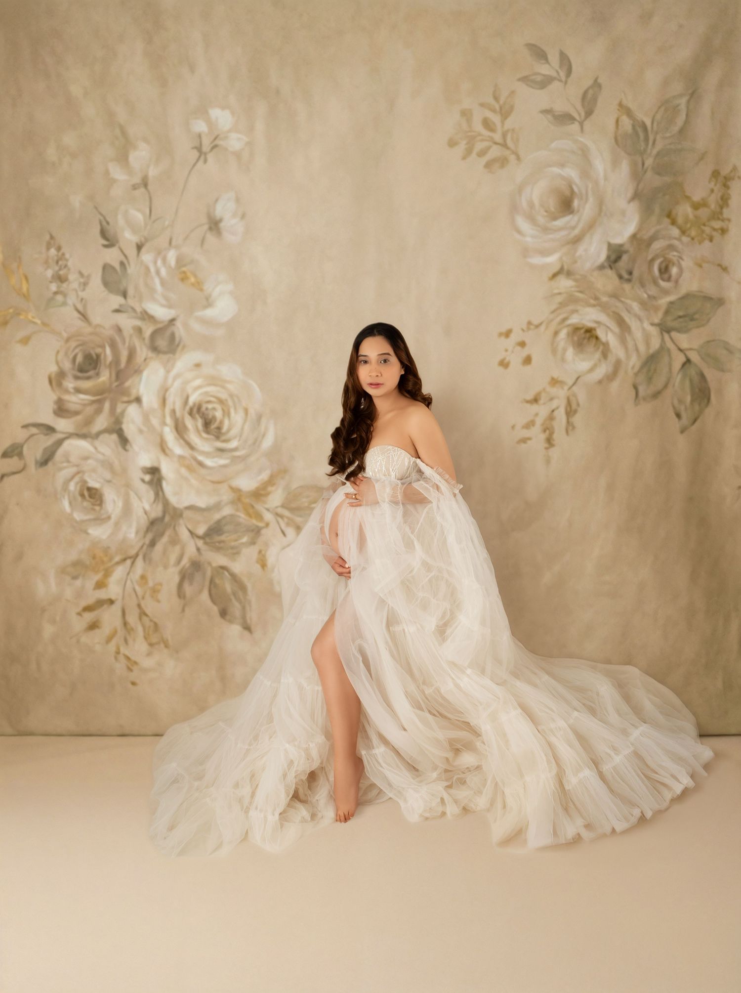 Elegant woman in flowing white tulle maternity gown sits before a golden floral backdrop.
