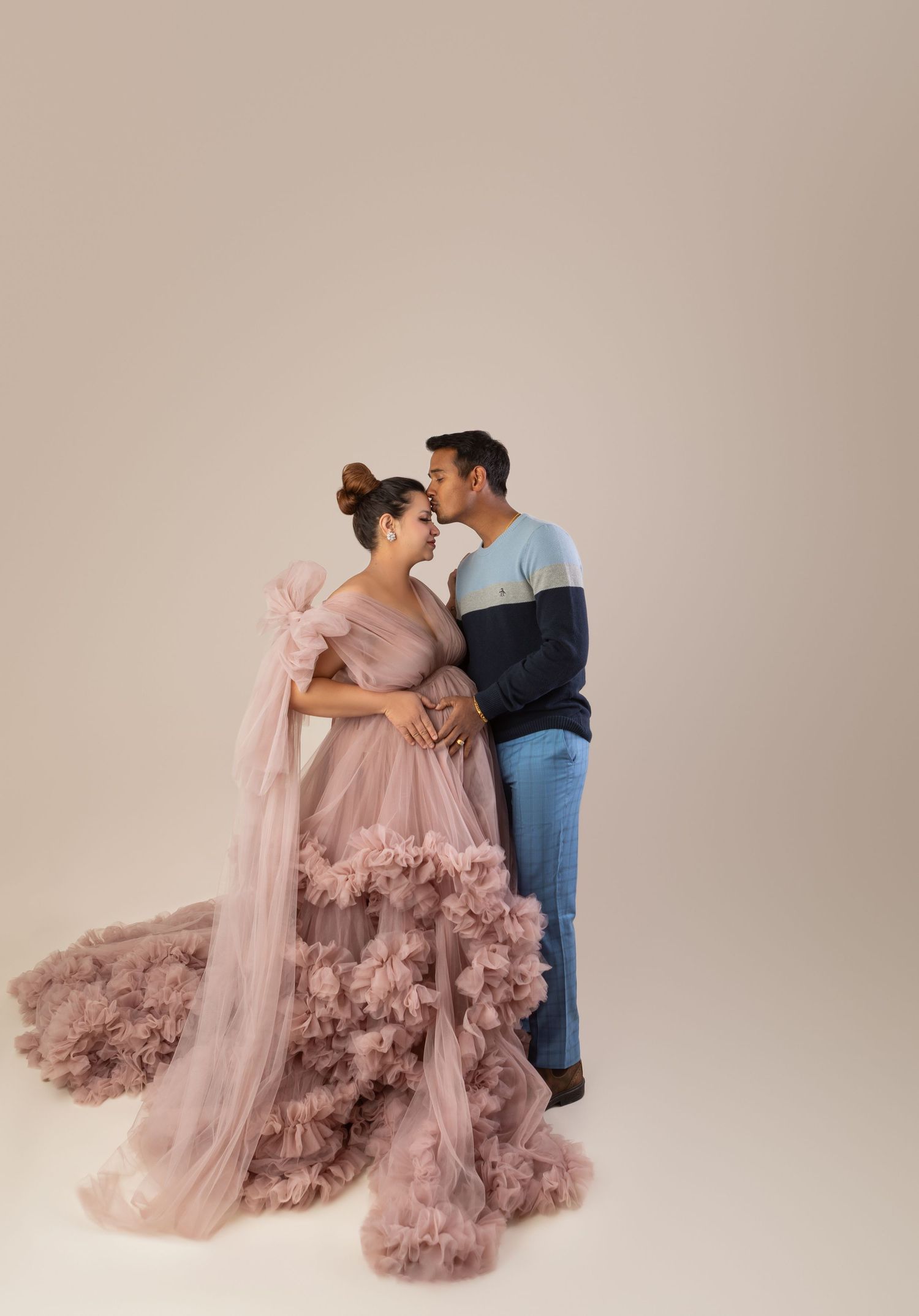 Expecting couple sharing tender moment, woman in dusty pink ruffled maternity gown during studio photoshoot.