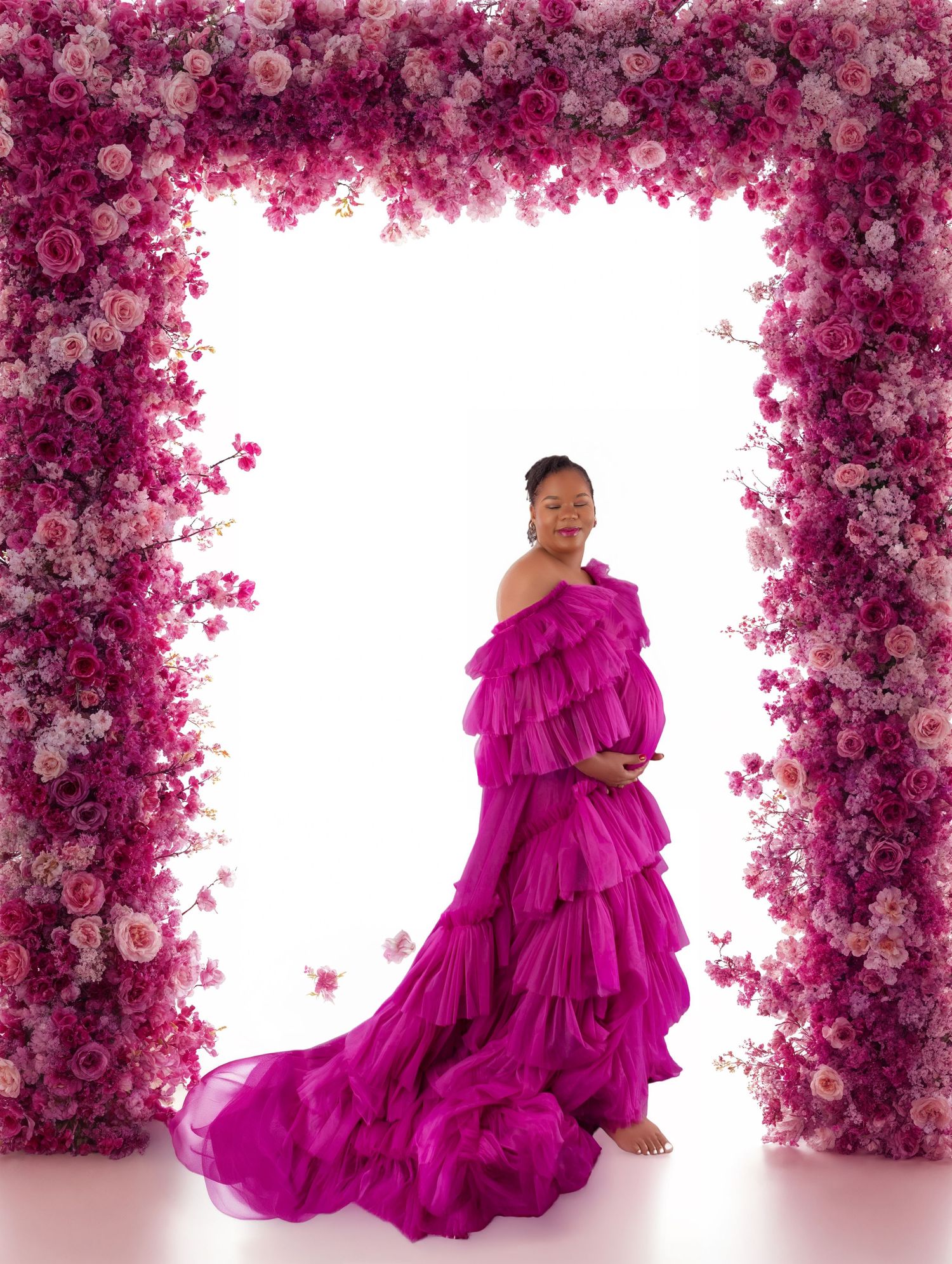 Pregnant woman in a ruffled fuchsia gown poses under a floral arch of pink and magenta roses for a maternity photo.