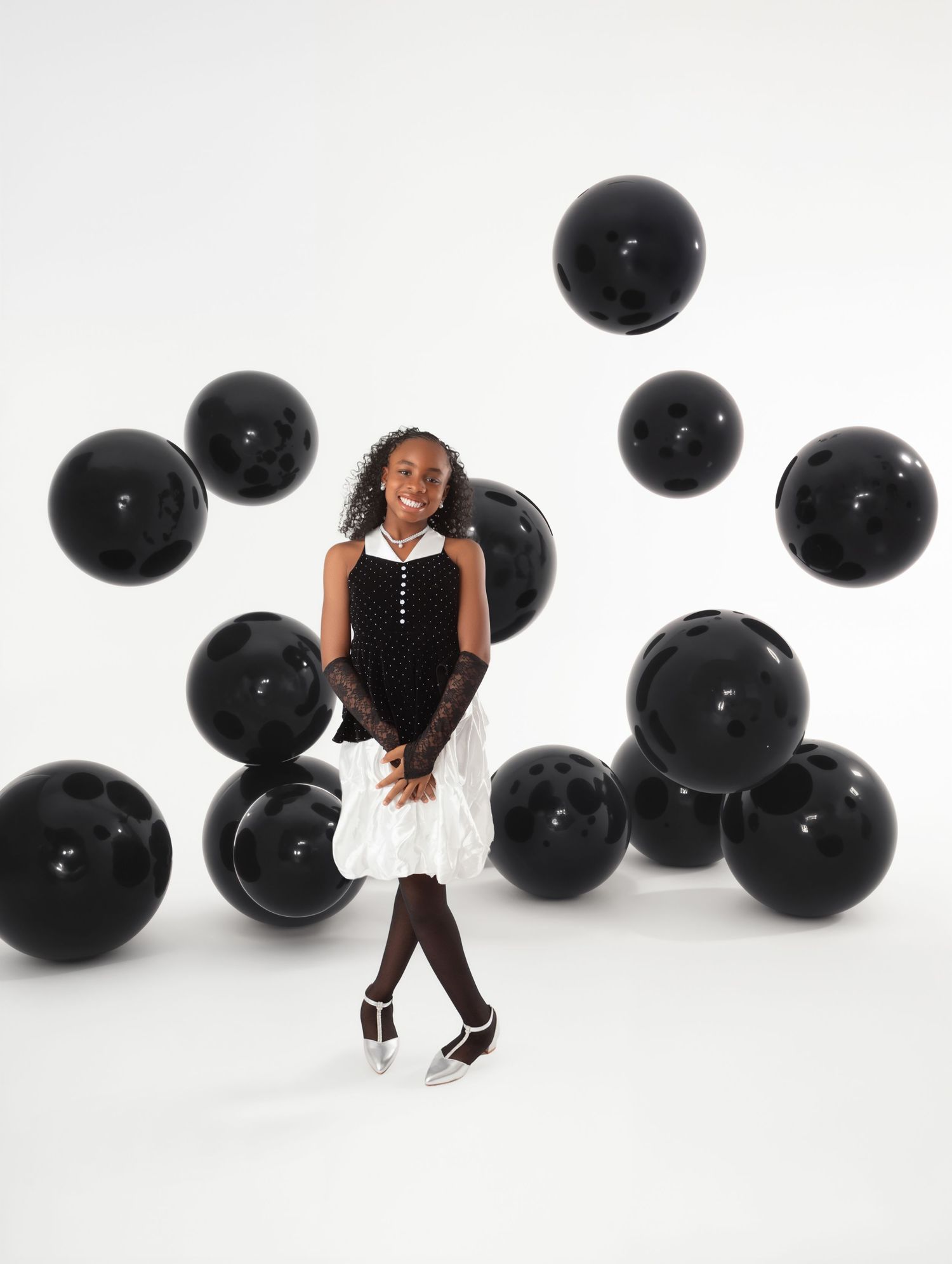 Young girl in black and white dress smiling among floating black balloons against white background.