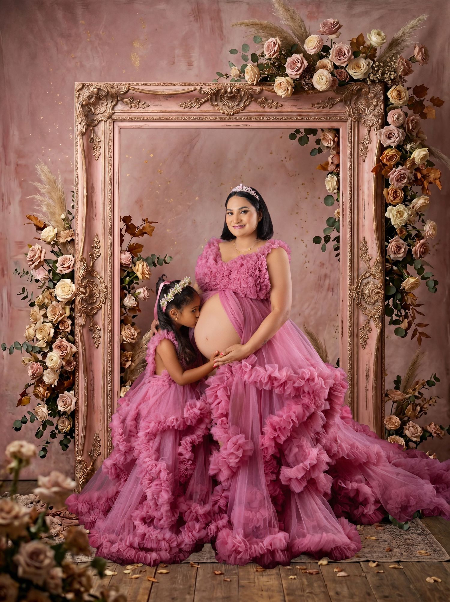Pregnant woman and young girl in matching pink ruffle gowns pose before a floral-adorned ornate frame backdrop.